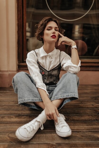 stylish-woman-jeans-white-sneakers-blouse-with-lace-sitting-floor-street-modern-woman-with-short-hair-poses-outside_197531-19313
